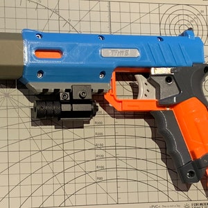 May include: A blue, gray, and orange toy gun with the word "Time" printed on the side. The toy gun has a black laser sight attached to the bottom and is resting on a grid surface.