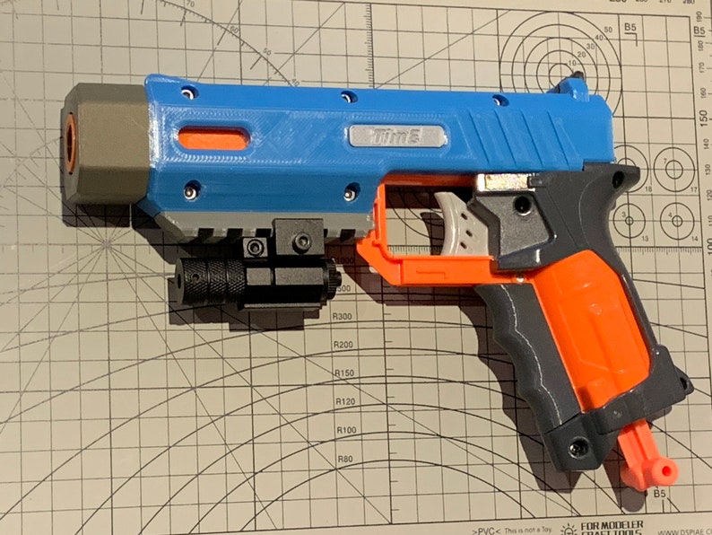 May include: A blue, gray, and orange toy gun with the word "Time" printed on the side. The toy gun has a black laser sight attached to the bottom and is resting on a grid surface.