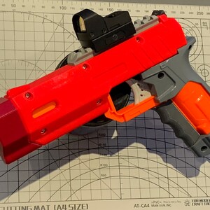 May include: A red and gray toy pistol with an attached black sight. The pistol has an orange barrel and grip, and is resting on a cutting mat with measurement markings.