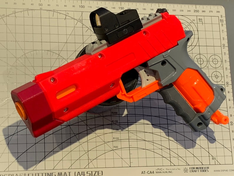 May include: A red and gray toy pistol with an attached black sight. The pistol has an orange barrel and grip, and is resting on a cutting mat with measurement markings.