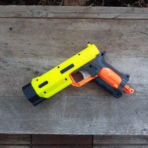 May include: A neon yellow and black toy gun with orange and gray accents. The toy gun is lying on a weathered wooden surface. The gun has a textured surface and a black handle.