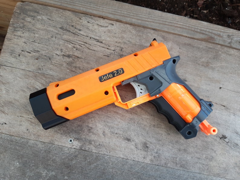 May include: An orange and gray toy gun with the text "Jefe 2.0" printed on the side. The toy gun is made of plastic and has a black barrel and handle. The toy gun is lying on a wooden surface.