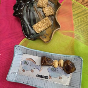 May include: A ceramic leaf-shaped bowl with sesame seed treats and a rectangular dish with fish illustrations, dates, and pistachios. The rectangular dish has the name "Edith P" printed on it. The background includes pink and green fabric.