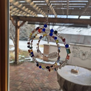 May include: A handcrafted spiral suncatcher with a copper wire frame, adorned with colorful glass beads. The beads are in various shapes and sizes, including clear, blue, red, and yellow. The suncatcher hangs from a metal hook, creating a decorative piece for a window or outdoor space.
