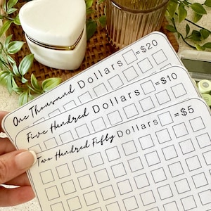 May include: Three white paper savings trackers with black text and grid boxes. The top sheet reads One Thousand Dollars with boxes equaling $20, the middle reads Five Hundred Dollars with boxes equaling $10, and the bottom reads Two Hundred Fifty Dollars with boxes equaling $5.
