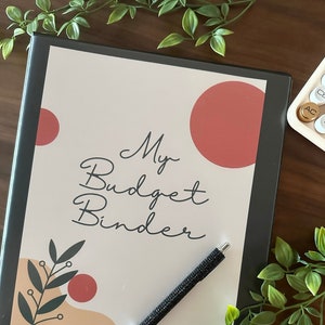 May include: A black binder with the text "My Budget Binder" on the front cover. The cover has a simple design with a red circle, a green leaf, and a tan background. A black pen with rhinestones is lying on the open binder.