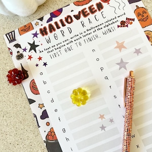 Könnte beinhalten: Ein Halloween Word Race-Spielblatt mit Halloween-Illustrationen. Ein roségoldener Stift und eine gelbe, blütenförmige Verzierung befinden sich auf dem Blatt. Der Text lautet: "Schreibe so schnell du kannst ein Wort, das mit Halloween zu tun hat und mit jedem Buchstaben des Alphabets beginnt. DER ERSTE, DER FERTIG IST, GEWINNT!"