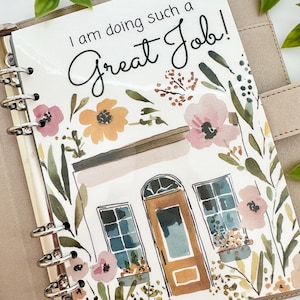 May include: A ring-bound planner with a beige cover featuring a watercolor illustration of a house surrounded by flowers and the text I am doing such a Great Job! The design includes a brown door, blue windows, and pink and yellow floral accents.