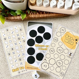 May include: Three game cards with a bee theme, featuring dice, circles, and hexagons. The cards have text that reads "Roll To" and "Bee Wild". A small die with a bee graphic is also present. The cards are on a light-colored surface.