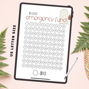 May include: A digital tablet displays a savings tracker for a $1,000 emergency fund. The design features a grid of 100 circles, each representing $10. Text includes "$1,000 emergency fund" and "US LETTER SIZE". A stylus and a cup of coffee are also visible.