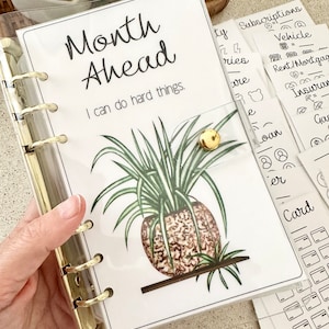 May include: A clear plastic binder with gold rings, a white page with the text "Month Ahead" and "I can do hard things." and a drawing of a potted plant. The binder has several tabs with handwritten text and icons for "Subscriptions", "Vehicle", "Rent/Mortgage", "Insurance", "Gas", "Loan", "Card".