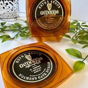 Peut inclure: Deux dessous de verre Guinness de couleur ambre avec le logo et le texte Guinness. Les dessous de verre sont carr&eacute;s avec des coins arrondis et une indentation circulaire. Le texte sur les dessous de verre indique "Extra Stout Guinness" et "St James's Gate Dublin".