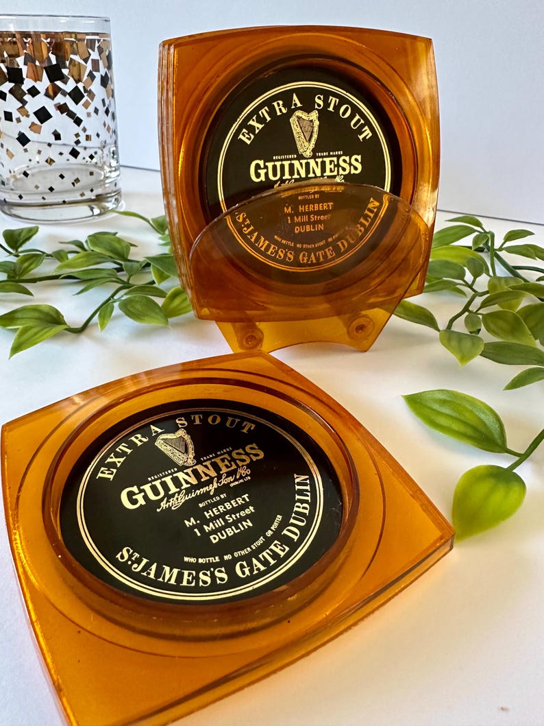Peut inclure: Deux dessous de verre Guinness de couleur ambre avec le logo et le texte Guinness. Les dessous de verre sont carr&eacute;s avec des coins arrondis et une indentation circulaire. Le texte sur les dessous de verre indique "Extra Stout Guinness" et "St James's Gate Dublin".