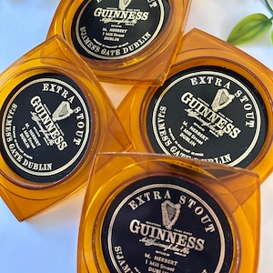 Peut inclure: Quatre dessous de verre carr&eacute;s Guinness de couleur ambre. Chaque dessous de verre pr&eacute;sente une &eacute;tiquette noire et dor&eacute;e avec le logo Guinness, les mots "Extra Stout" et l'adresse "St. James's Gate Dublin."