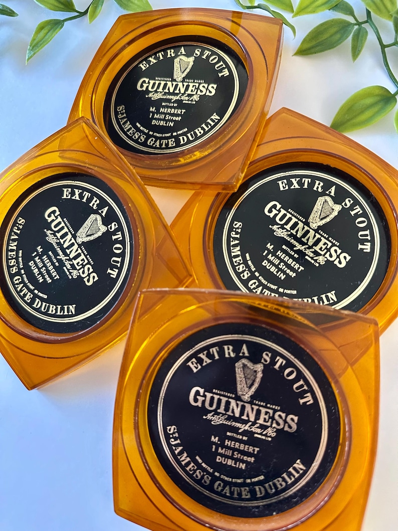 Peut inclure: Quatre dessous de verre carr&eacute;s Guinness de couleur ambre. Chaque dessous de verre pr&eacute;sente une &eacute;tiquette noire et dor&eacute;e avec le logo Guinness, les mots "Extra Stout" et l'adresse "St. James's Gate Dublin."