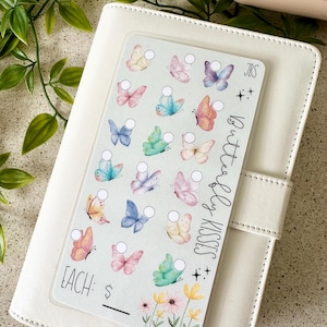 May include: A rectangular sticker sheet featuring twenty-one watercolour-style butterflies in various pastel colours, each paired with a small white circular checkbox. The text reads Butterfly Kisses, JBS, and EACH: $. Three small flowers are at the bottom.