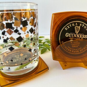 Peut inclure: Un verre transparent orn&eacute; de motifs carr&eacute;s dor&eacute;s, noirs et cuivr&eacute;s, pos&eacute; sur un dessous de verre orange, &agrave; c&ocirc;t&eacute; d'un ensemble de dessous de verre Guinness dans un support orange. Les dessous de verre pr&eacute;sentent le logo Guinness.