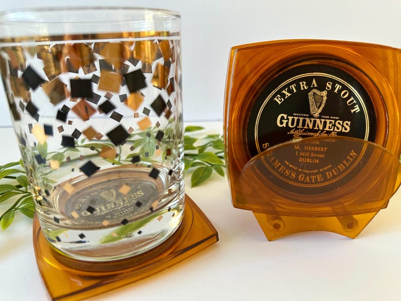 Peut inclure: Un verre transparent orn&eacute; de motifs carr&eacute;s dor&eacute;s, noirs et cuivr&eacute;s, pos&eacute; sur un dessous de verre orange, &agrave; c&ocirc;t&eacute; d'un ensemble de dessous de verre Guinness dans un support orange. Les dessous de verre pr&eacute;sentent le logo Guinness.