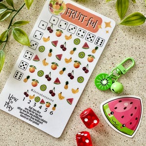 May include: A white card with the text "FRUIT-ful" and a grid of fruit icons. The card is next to two red dice and a green watermelon sticker with a smiling face. The card has instructions for a game called "How to Play" with the text "Roll two dice, one die for top row, other one for descending column. Save where they intersect based on the symbol."