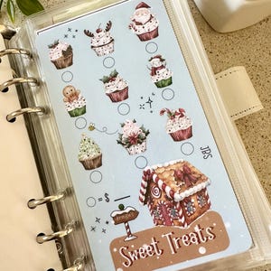 Puede incluir: Una hoja de seguimiento azul claro para archivador de anillas con ilustraciones de cupcakes navideños, una casa de jengibre y un cartel que dice Kringle's. El texto Sweet Treats aparece en la parte inferior junto a círculos de control.
