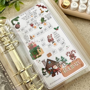 Puede incluir: Un rastreador de ahorro con temática navideña en una funda de plástico transparente. Presenta ilustraciones navideñas, incluyendo a Papá Noel, regalos y árboles. La palabra "HOLIDAZE" es visible.