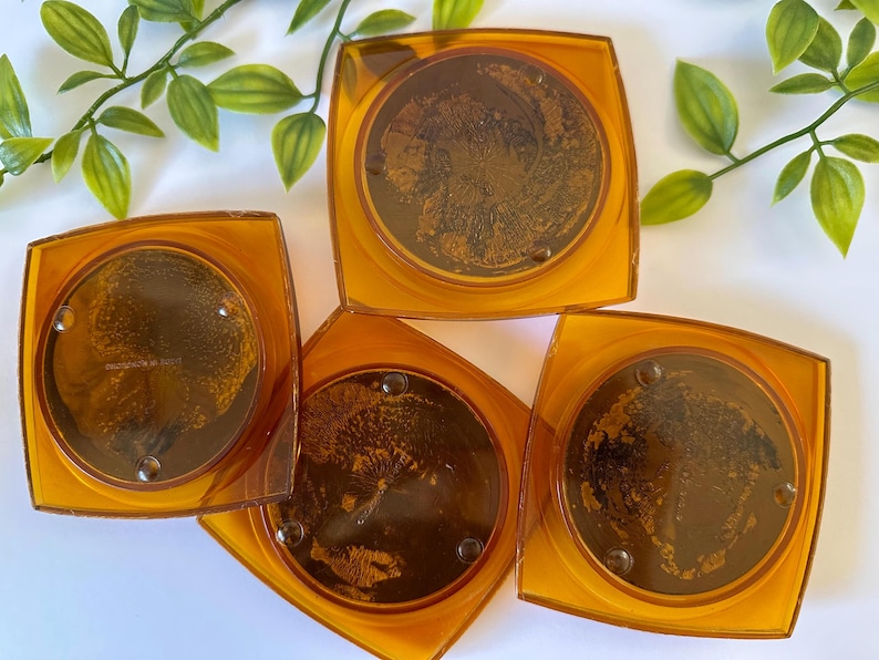 Peut inclure: Quatre dessous de verre carr&eacute;s ambr&eacute;s avec un motif circulaire au centre. Les dessous de verre ont un bord transparent et un centre brun fonc&eacute;, peut-&ecirc;tre &agrave; motifs. Ils sont dispos&eacute;s sur une surface blanche avec des brins de feuilles vertes.