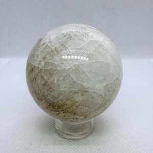 May include: A clear quartz sphere with internal inclusions, resting on a clear acrylic stand. The sphere has a polished surface with a mix of clear and light brown tones. The quartz appears to be translucent, allowing light to pass through.