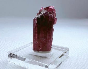 May include: A deep pink, multifaceted, elongated crystal with a rough texture. The crystal is displayed on a clear acrylic base.