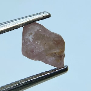 May include: A pink, faceted, rough, natural gemstone held by tweezers. The gemstone is translucent and has a slight yellow hue.