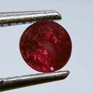 Op de afbeelding: Een dieprode robijn edelsteen, rond van vorm, wordt vastgehouden door een pincet. De robijn heeft een licht troebele uitstraling met een vleugje glans.