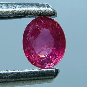 May include: A vibrant pink oval-shaped ruby gemstone held in tweezers. The ruby has a faceted cut and is translucent.