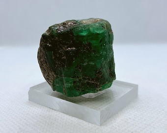 Può includere: Un grande cristallo di smeraldo verde grezzo con un colore verde scuro. Il cristallo è su un supporto di esposizione in vetro trasparente.