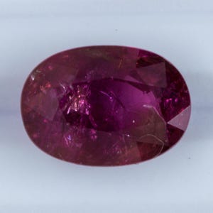 Könnte beinhalten: Ein oval geschliffener Rubin-Edelstein, der einen tiefroten Farbton mit inneren Einschlüssen aufweist. Der Edelstein ist poliert und reflektiert das Licht über seine Oberfläche. Die Form ist länglich, geeignet für Schmuck.