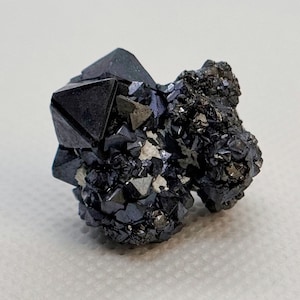 May include: A cluster of black and iridescent pyritohedral crystals, with a metallic sheen. The crystals are clustered together in a rough, irregular shape.