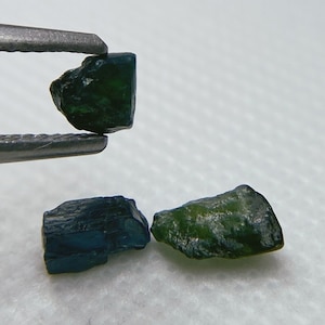 May include: Three rough, dark green gemstones. The gemstones are various shapes and sizes. The gemstones are being held by tweezers.