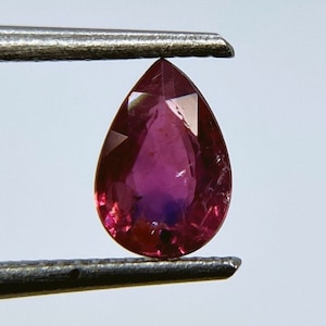 May include: A pear-shaped ruby gemstone with a deep red hue and a slight purple tint. The gemstone is held in tweezers.