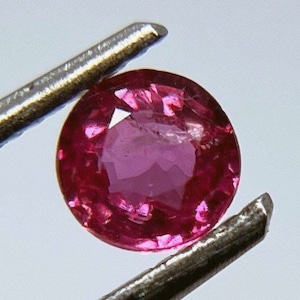 May include: A round, pink gemstone with a faceted cut. The gemstone is held in place by a metal tweezer.