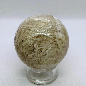 May include: A clear, spherical crystal ball with a network of golden, needle-like inclusions. The sphere is displayed on a small, clear glass stand, showcasing the intricate internal patterns and textures of the crystal.