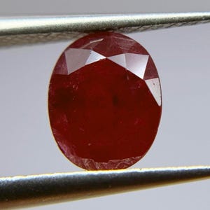 Puede incluir: Una piedra preciosa ovalada de color rojo oscuro con un corte facetado. La piedra preciosa está sujeta por unas pinzas.