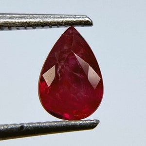 Pode incluir: Uma gema de rubi em forma de pêra com uma cor vermelha profunda. A gema é segurada em pinças e tem um corte facetado.