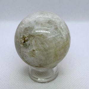 May include: A clear quartz crystal sphere with internal inclusions and a slight green hue. The sphere is displayed on a small, clear, round stand, showcasing its spherical shape and natural patterns.