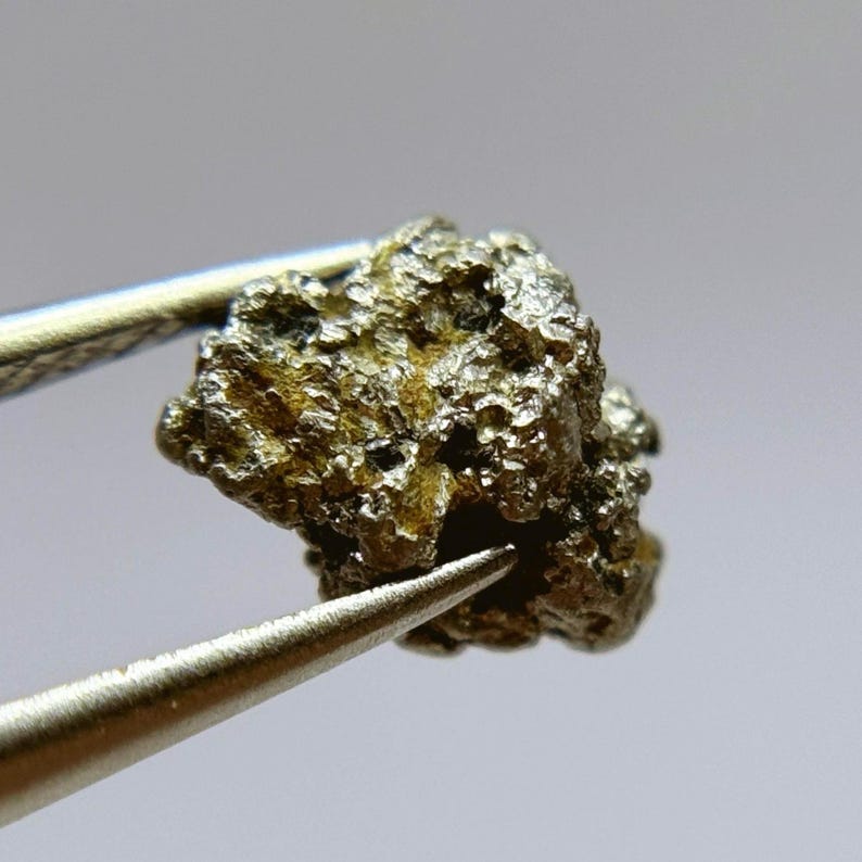 May include: A small, irregularly shaped piece of metallic silver-gray material, held by tweezers. The material has a rough, textured surface.