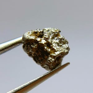 May include: A small, rough, metallic, silver-colored rock with a textured surface. The rock is being held by tweezers.