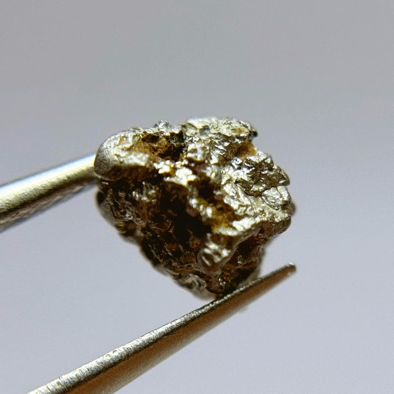 May include: A small, rough, metallic, silver-colored rock with a textured surface. The rock is being held by tweezers.