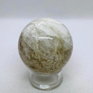 May include: A clear quartz sphere with internal inclusions, resting on a clear stand. The sphere has a mix of clear and light brown hues, with visible needle-like formations within the crystal. The overall effect is a natural, earthy aesthetic.