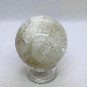 May include: A translucent, spherical crystal ball with a light green and white color scheme. The crystal ball is resting on a clear, circular stand. The surface of the sphere has a textured appearance.