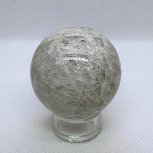 May include: A clear quartz crystal sphere, resting on a small, clear stand. The sphere has a cloudy, translucent appearance with internal veining and a polished surface. The background is a plain white.