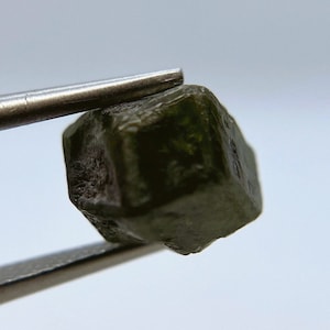 May include: A dark green, faceted gemstone with a cubic shape. The gemstone is held by tweezers.