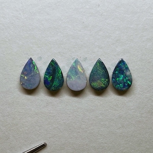 May include: Five teardrop-shaped opal gemstones in various shades of green and white. The opals are arranged in a row on a white surface.