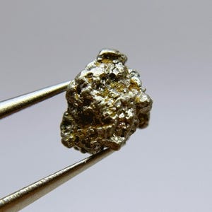 May include: A small, rough, silver-colored nugget of platinum. The nugget is being held by tweezers against a white background.